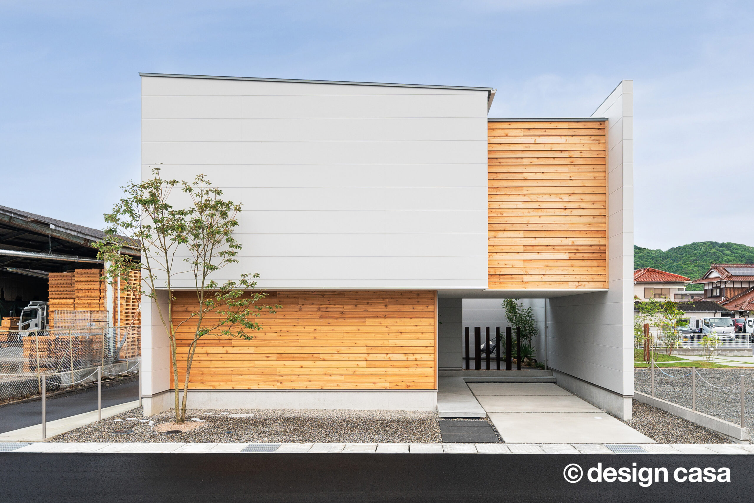 白い外壁と杉板張りを組み合わせた、箱型の外観住宅。玄関前の植栽と砂利のアプローチが見える