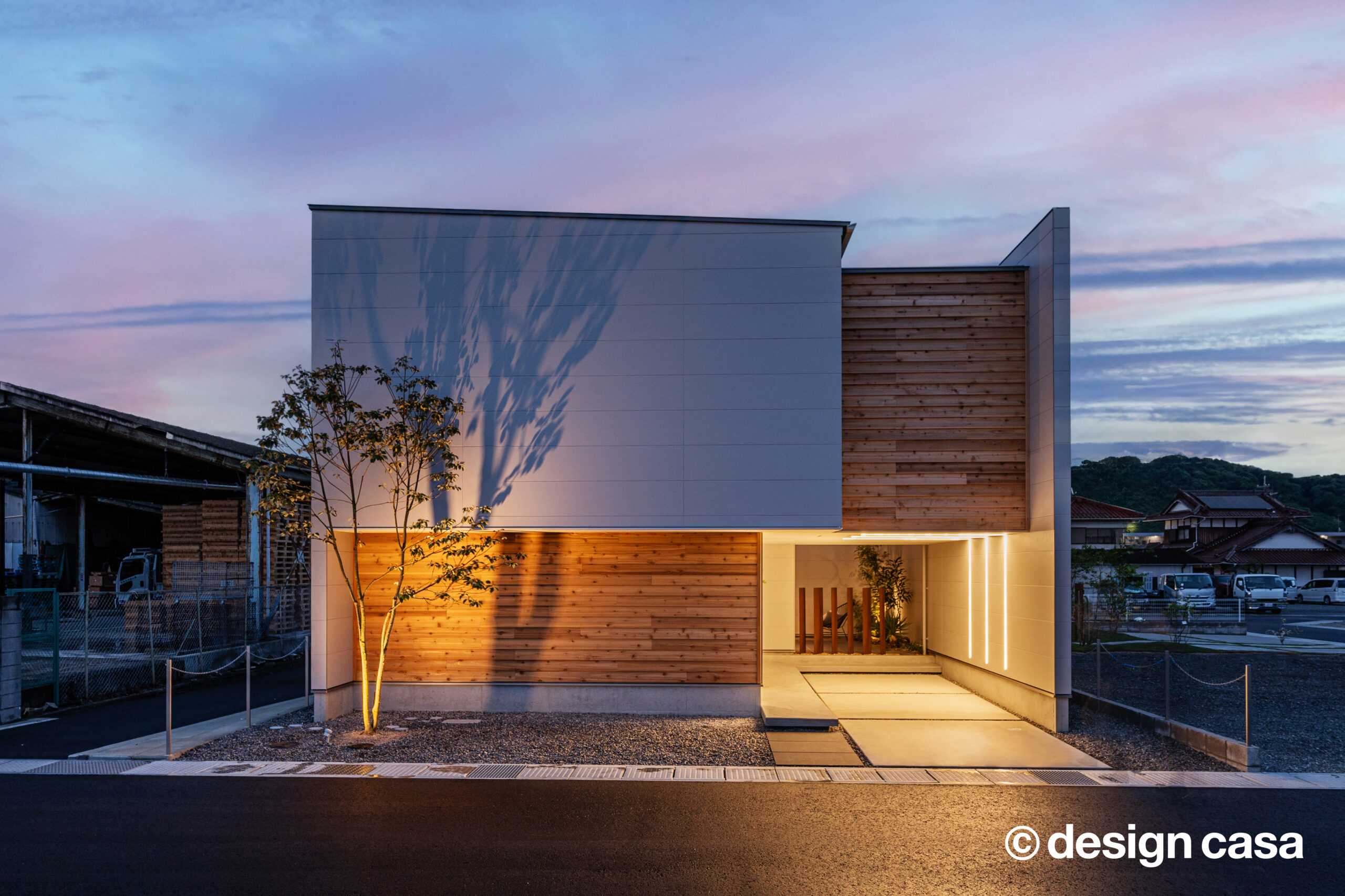 夕暮れの空の下、杉板張りの外壁と白い壁の外観住宅。ライトアップされた木壁とアプローチが浮かび上がる