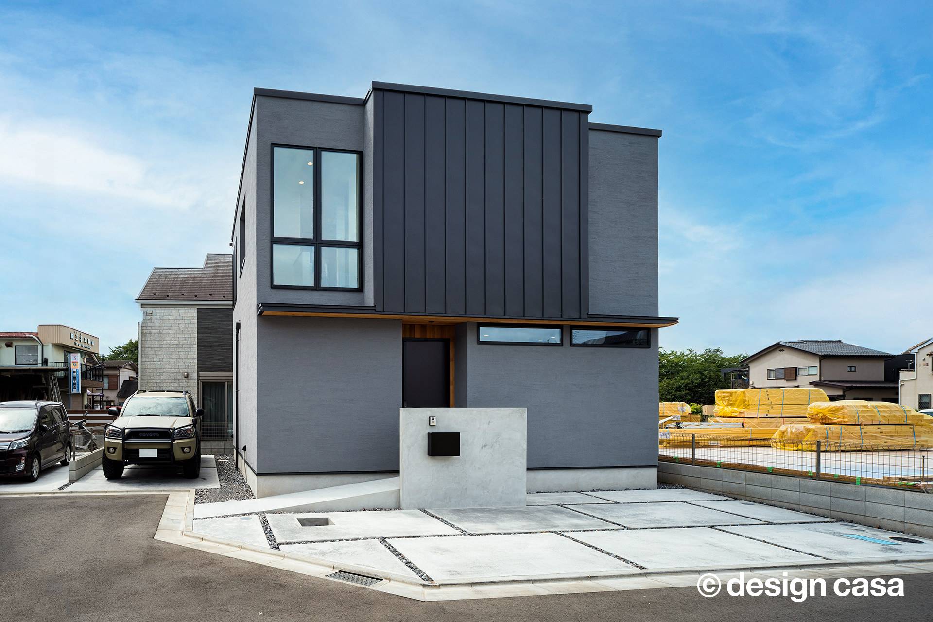グレーの外壁に黒いガルバリウムの縦張りを組み合わせた、2階建ての外観住宅。玄関前には表札付きのコンクリート壁がある
